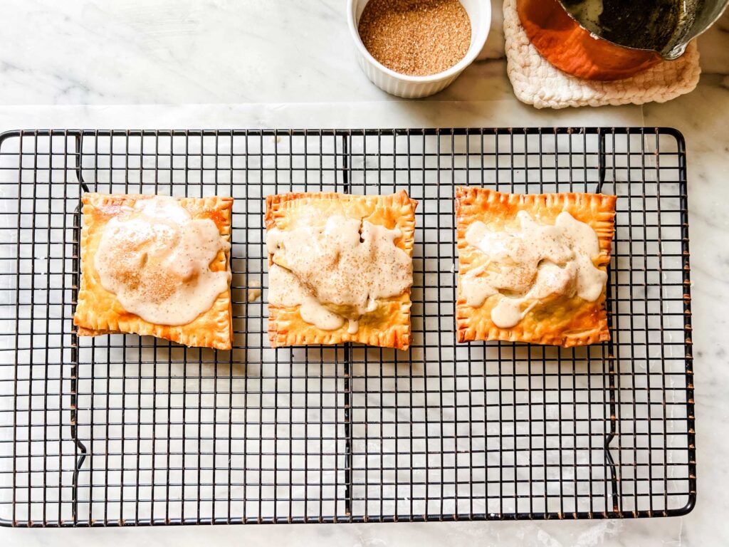 Three apple pie pop tarts with a brown butter glaze rest on a wire cooling rack.