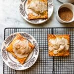 Three apple pie pop tarts. Two are on small floral plates and one is resting on a wire cooling rack. Turbinado surgar is in a small, white ramekin.