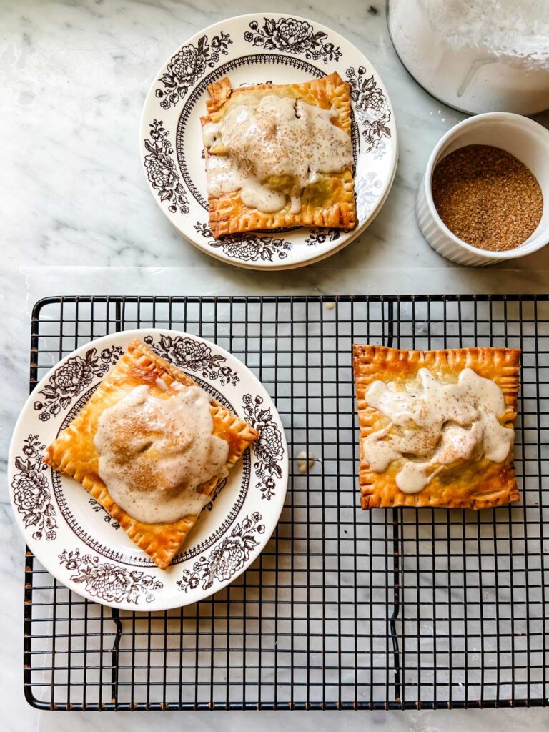 Three apple pie pop tarts. Two are on small floral plates and one is resting on a wire cooling rack. Turbinado surgar is in a small, white ramekin.