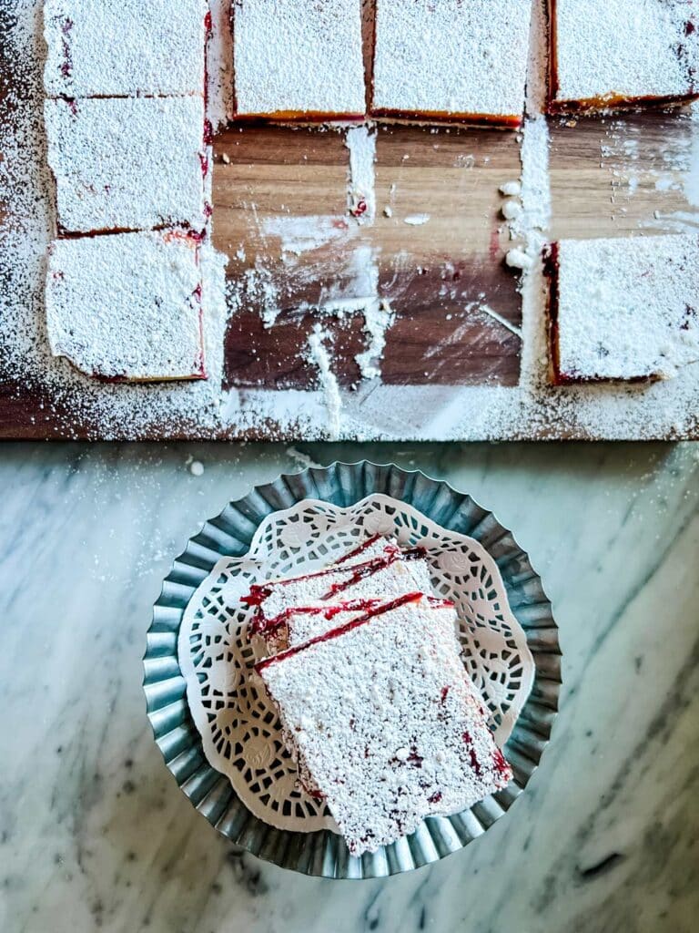 Some cranberry and lemon bars are on a cutting board and have been topped with confectioners' sugar. Other bars have been stacked on a cake stand.