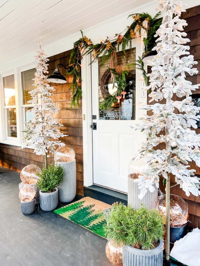 Two white Christmas trees flank each side of the front door. The door is draped with garland at the top. Magical light bulbs all around give the port a magical glow.