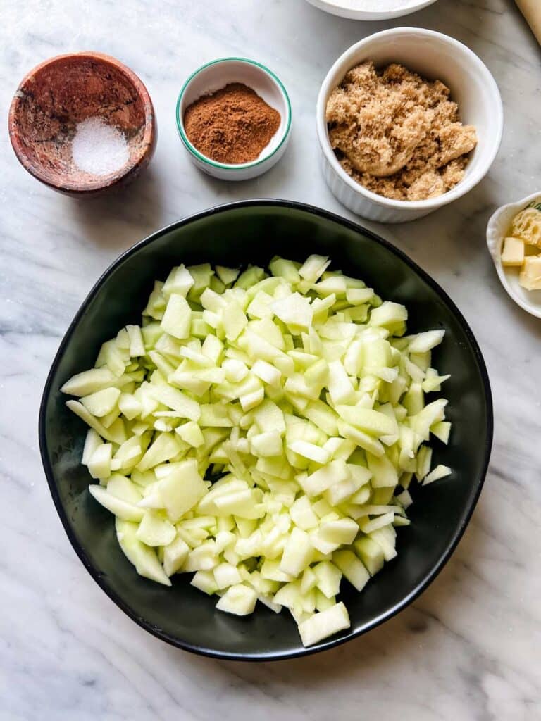 Diced apples are in a large black bowl. In small bowls are butter and brown sugar, salt, butter and bread cubes, and turbinato sugar.