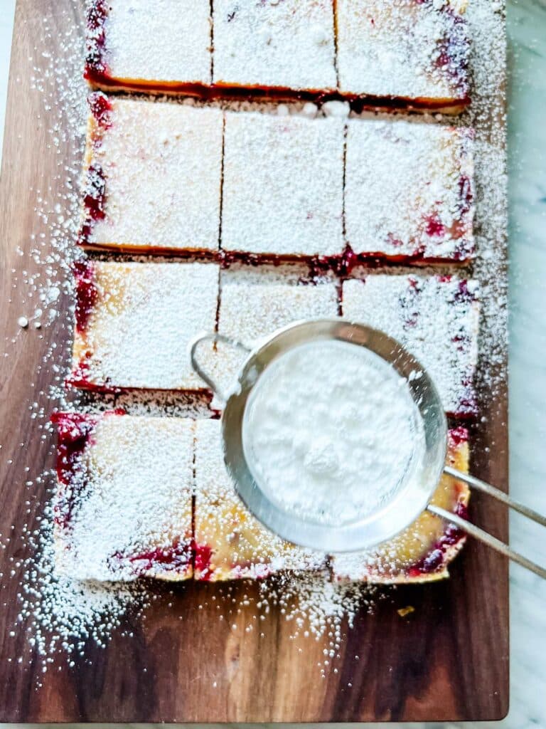 Cranberry and lemon bars have been cut into individual servings. They are being dusted with confectioners' sugar.