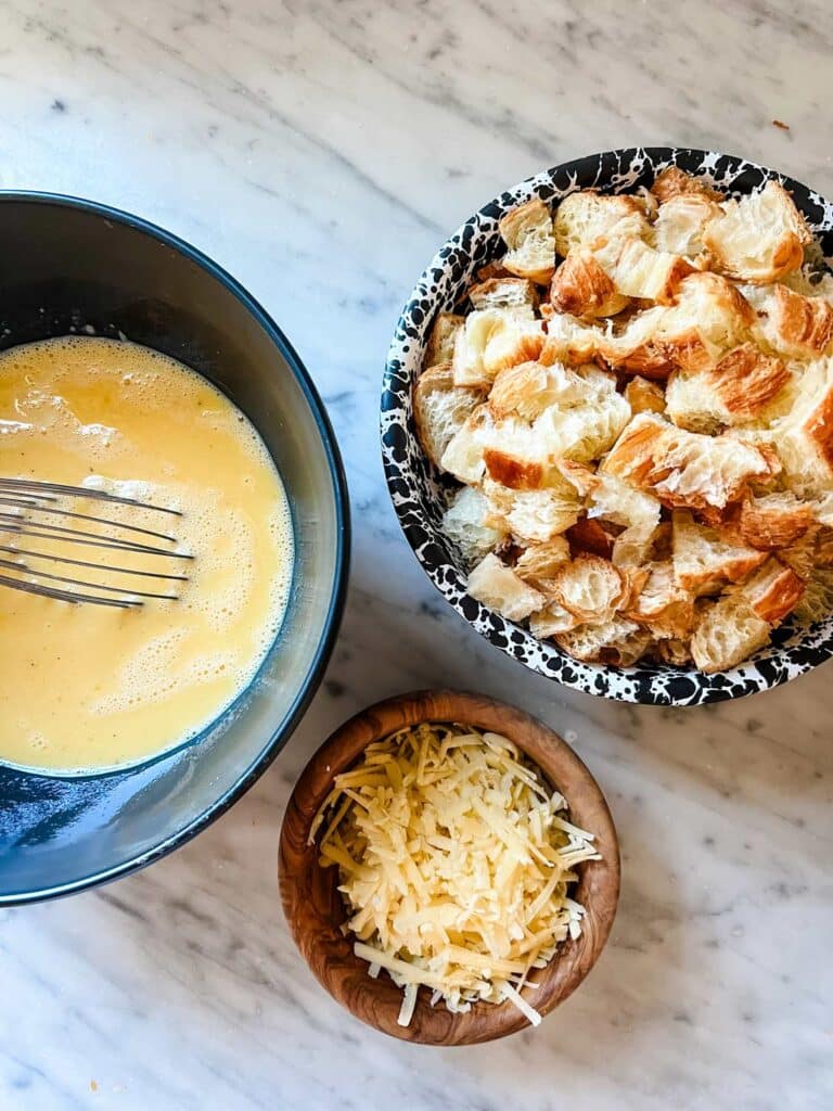 Croissants in a bowl have been diced, eggs and milk have been whisked and gruyere cheese has been grated.