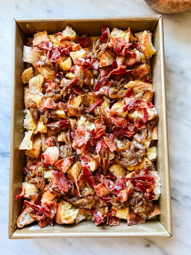 Diced croissants are topped with caramelized onions and chopped bacon. They are in a baking pan.