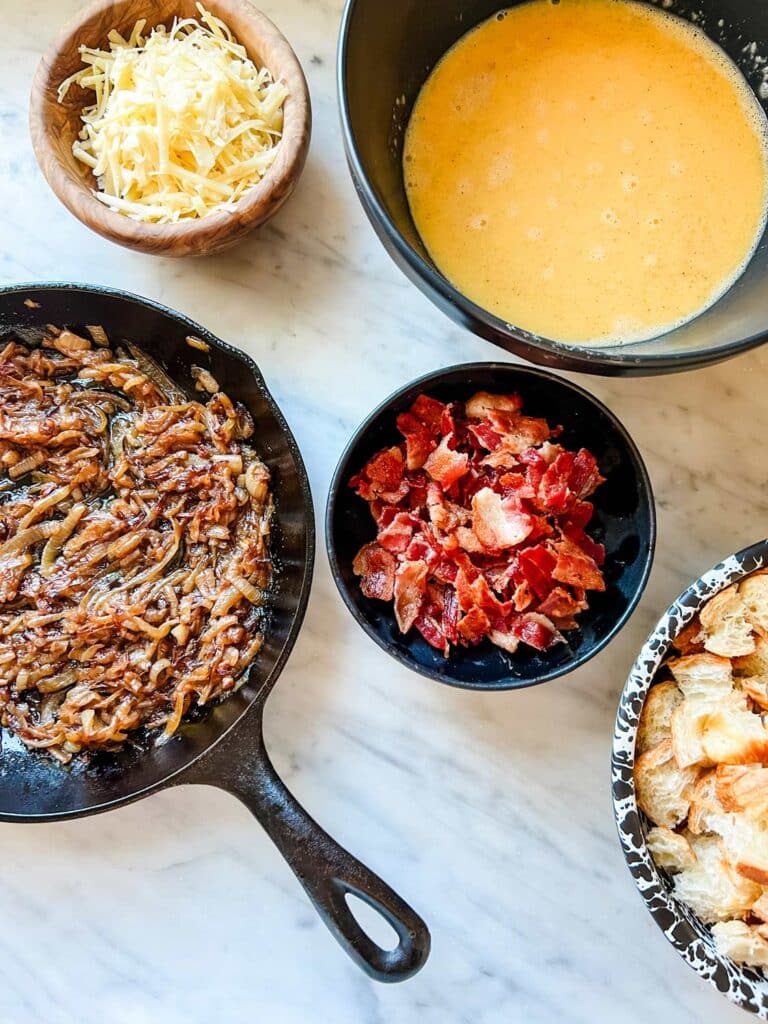 Onions are caramelized in a Lodge skillet, eggs and milk have been whisked, bacon has been cooked and chopped and the croissants have been diced into bite-sized pieces.