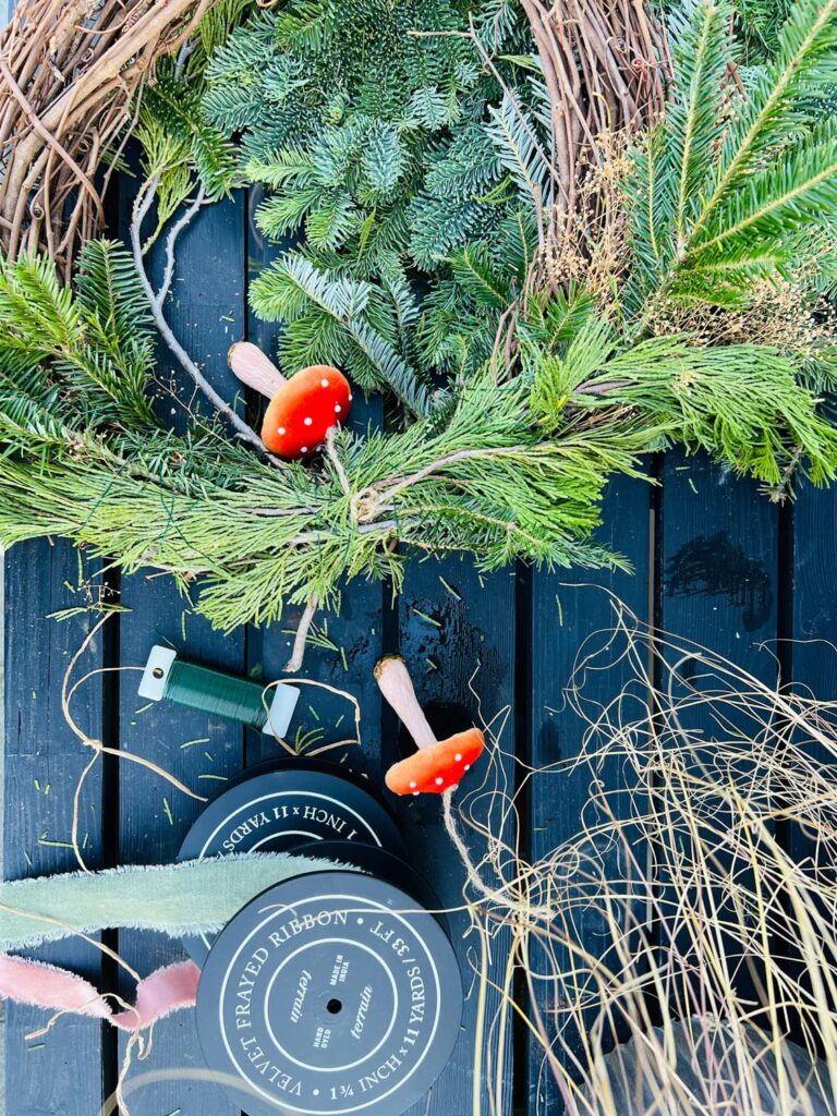 A wreath form, wrapped with fresh greenery lay on a black work bench. Ribbon and mushroom ornaments provided the decorations.
