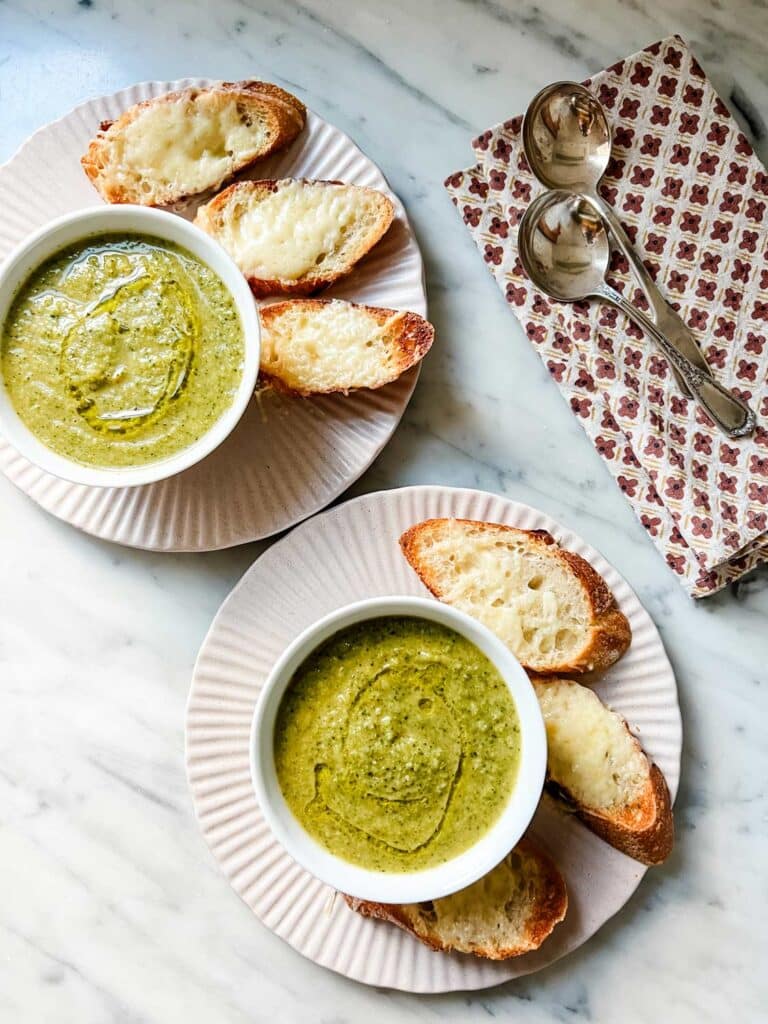 Broccoli soup is served in small white bowls on pink plates with slices of toasted baguettes topped with gruyere cheese.