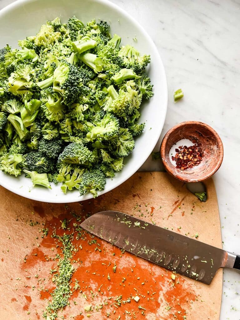 Broccoli florets in a white bowl next to a cutting board and knife.