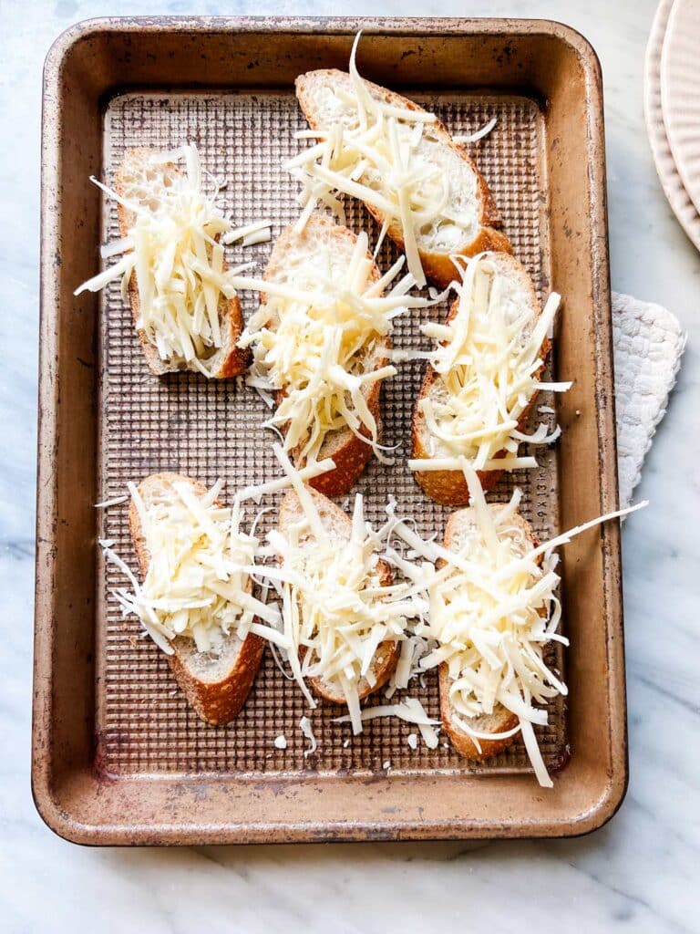Baguette slices are on a baking sheet and topped with grated gruyere cheese and are ready for toasting.