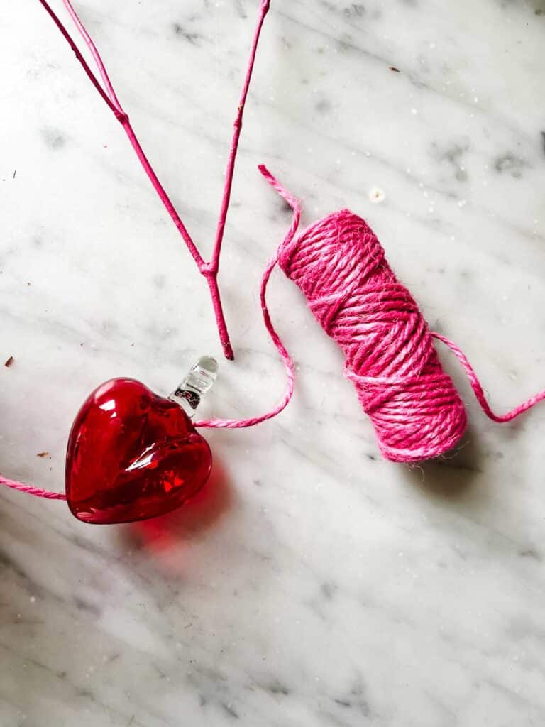 Pink twine, a twig painted pink and a glass heart ornament on marble 