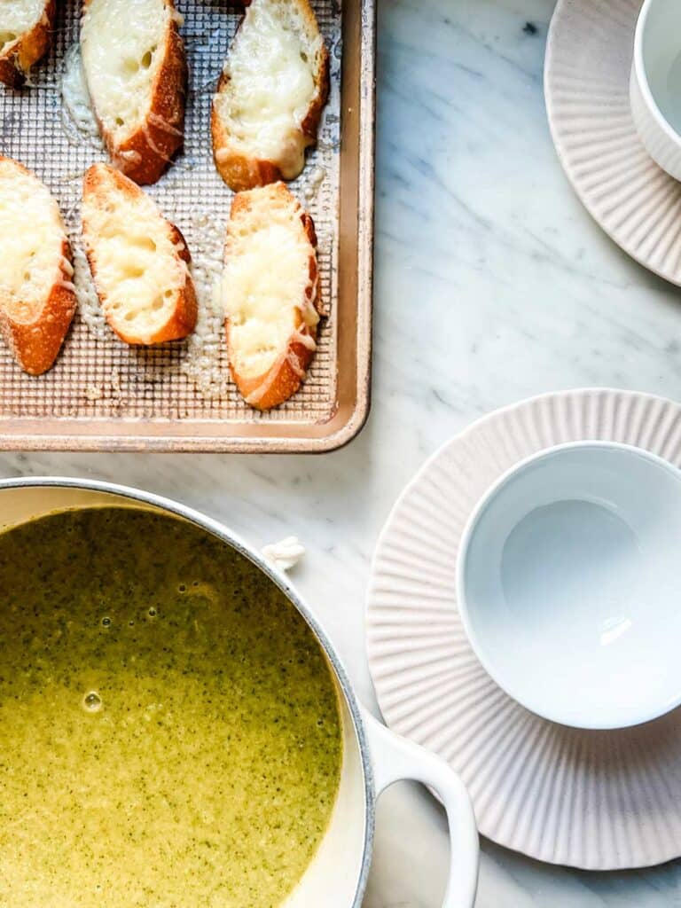 Broccoli soup in a Dutch oven sits next to toasted baguette slices and white bowls on pink plates.