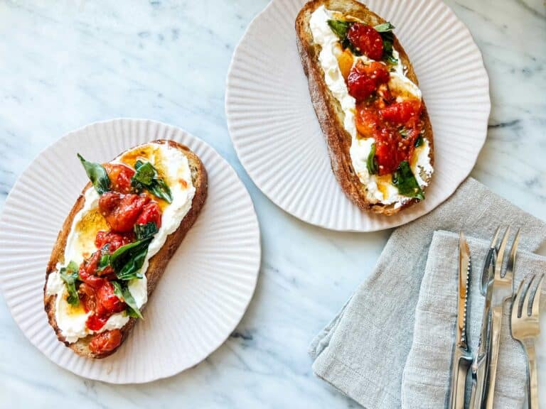 Garlicky ricotta cheese and tomato confit tartines are served on small pink plates with a scallop design. Linen napkins are next to the plates.