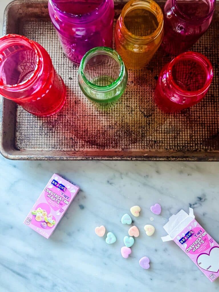Tinted and baked glass jars are cooling on a cookie sheet. Candy conversation hearts are in the foreground.