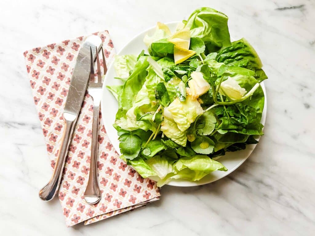 Via Carota's insalata verde is maybe the best salad you'll ever have. It's served on a white plate here. A napkin, fork, and knife are sitting next to it and ready for you or a guest to eat.
