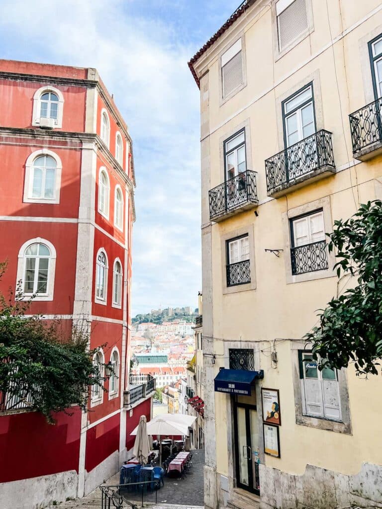 A view down a crooked narrow street in Lisbon. At the bottom sits a nice cafe with outdoor seating and tables.