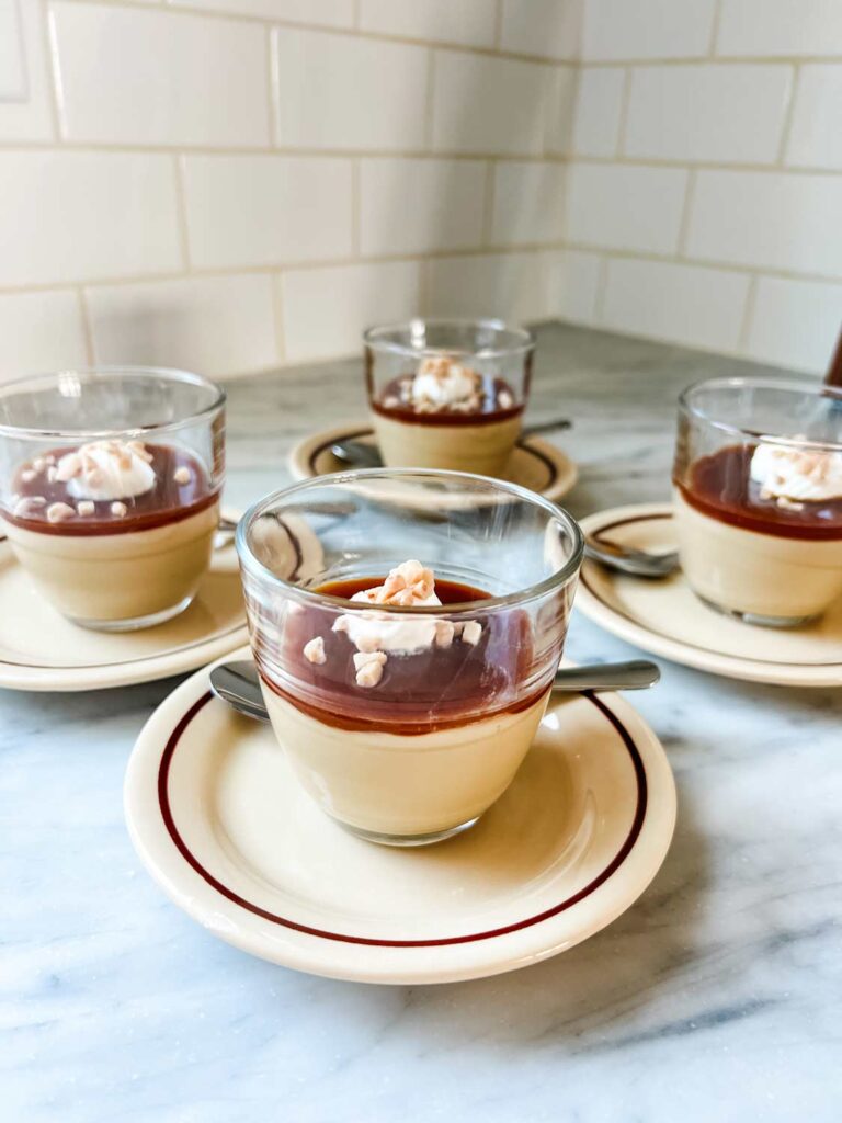 Homemade butterscotch pudding with salted caramel sauce, whipped cream, and heath bar crunch sprinkled on top are served in small French glasses.