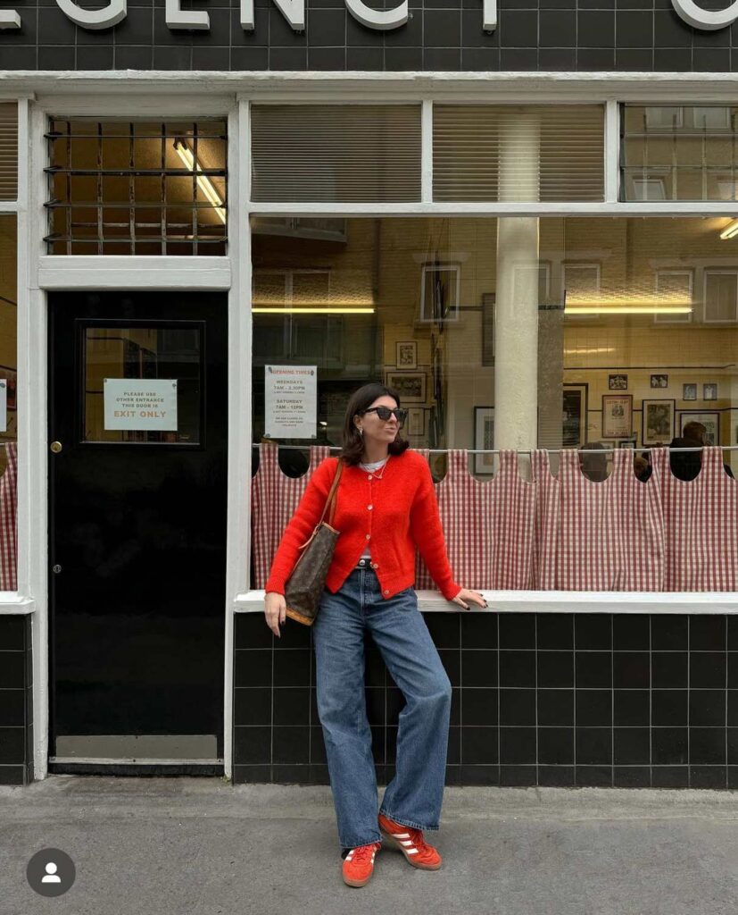 The digital creator, Francesca Saffari, leans against a building wearing red Adidas Gazelle sneakers and a red cardigan sweater.