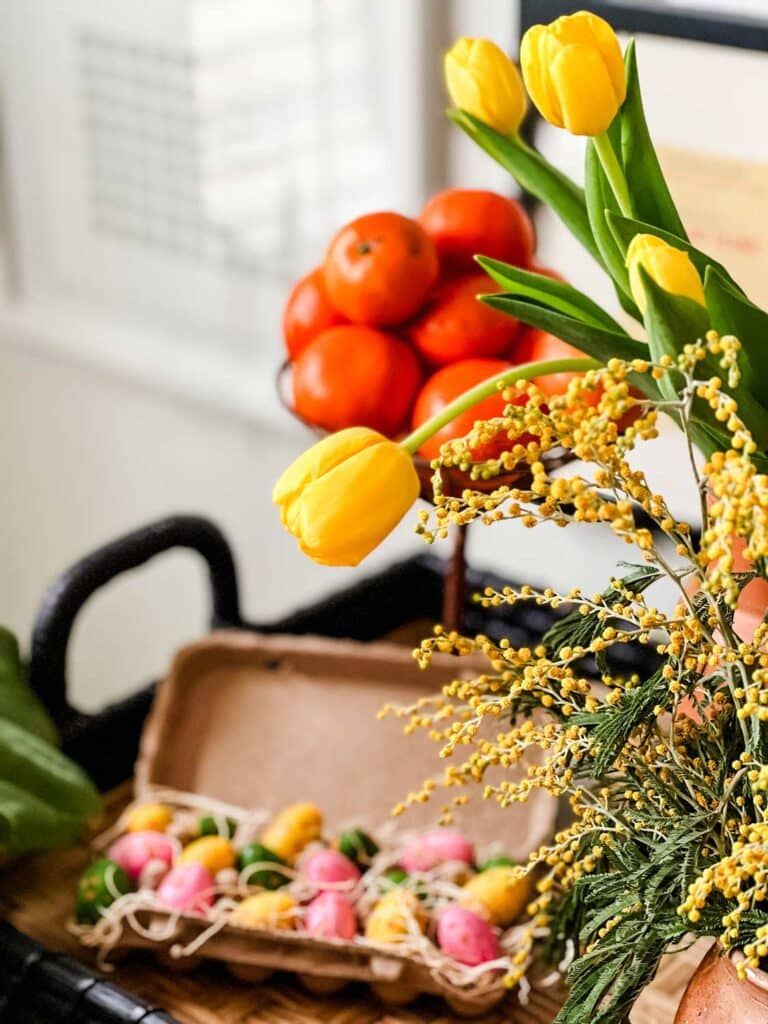 How to Set a Colorful Easter Table Setting 2024-chocolate covered Easter eggs in egg carton with tulips and mimosa, and mandarin in nest stand 