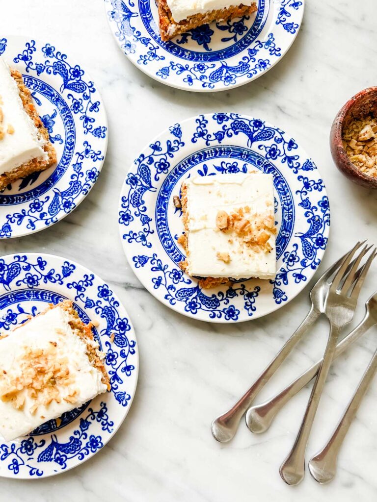 Cream cheese frosted carrot cake sprinkled with nuts is served on small blue and white French dessert plates.