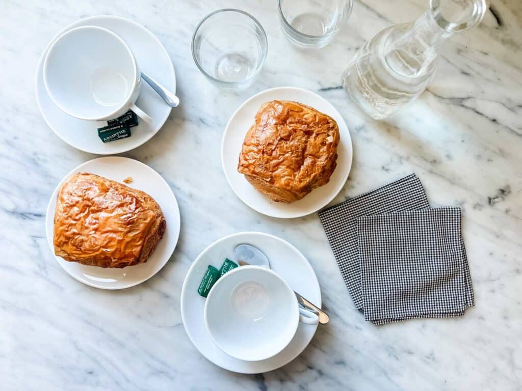 Two croissants on plates next to white Apilco coffee cups and saucers. Green French sugar cubs are also on the saucers and black and white napkins are next to them.