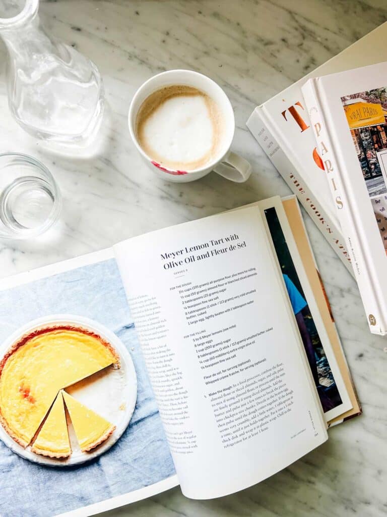 A french cookbook is on the kitchen counter next to a cup of coffee with steamed milk.