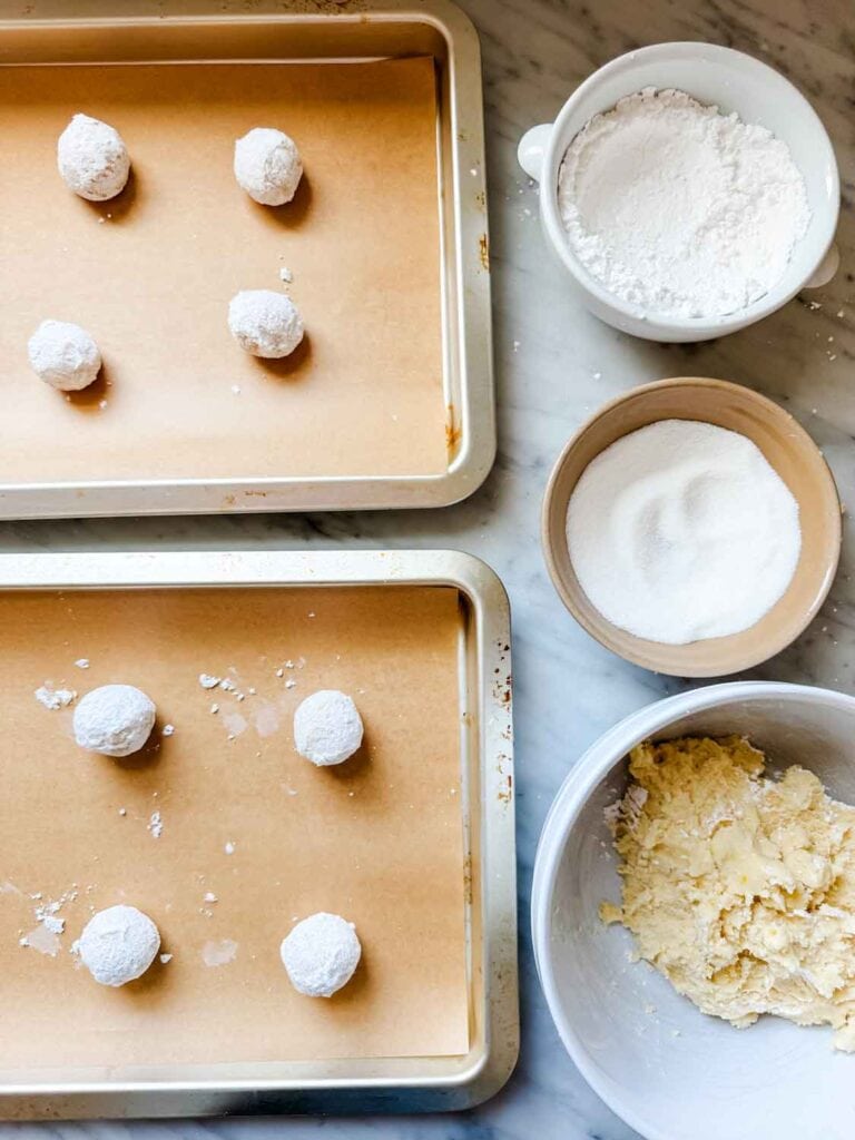 Cookie dough has been rolled into balls and rolled again in granulated sugar and powdered sugar, and placed on a parchment paper-lined baking sheet.