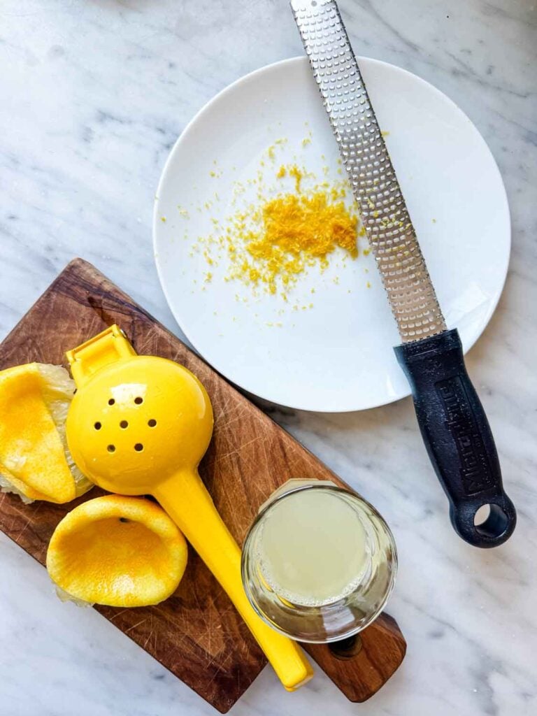 A juicer and juiced lemons are on a wooden cutting board next to a glass of freshly squeezed lemon juice. On a white plate is a microplane with zest from a couple of lemons.