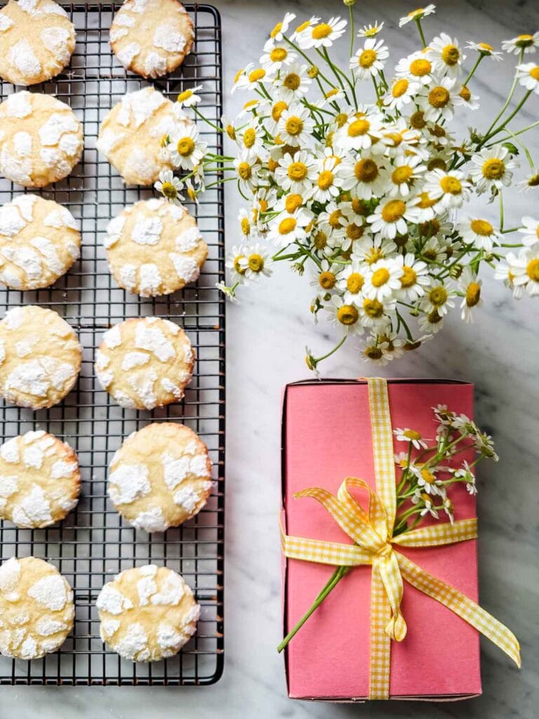 Lemon crinkle cookies are on a wire cooling rack next to a vase full of chamomile flowers and a pink box of cookies wrapped with yellow gingham ribbon and decorated with a small sprig of chamomile flowers.