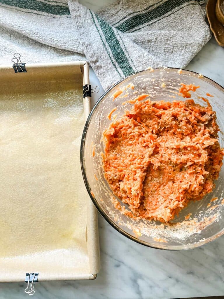 Wet and dry ingredients for low-carb carrot cake have been mixed in a large bowl. A cake pan lined with parchment paper sits next to the bowl.