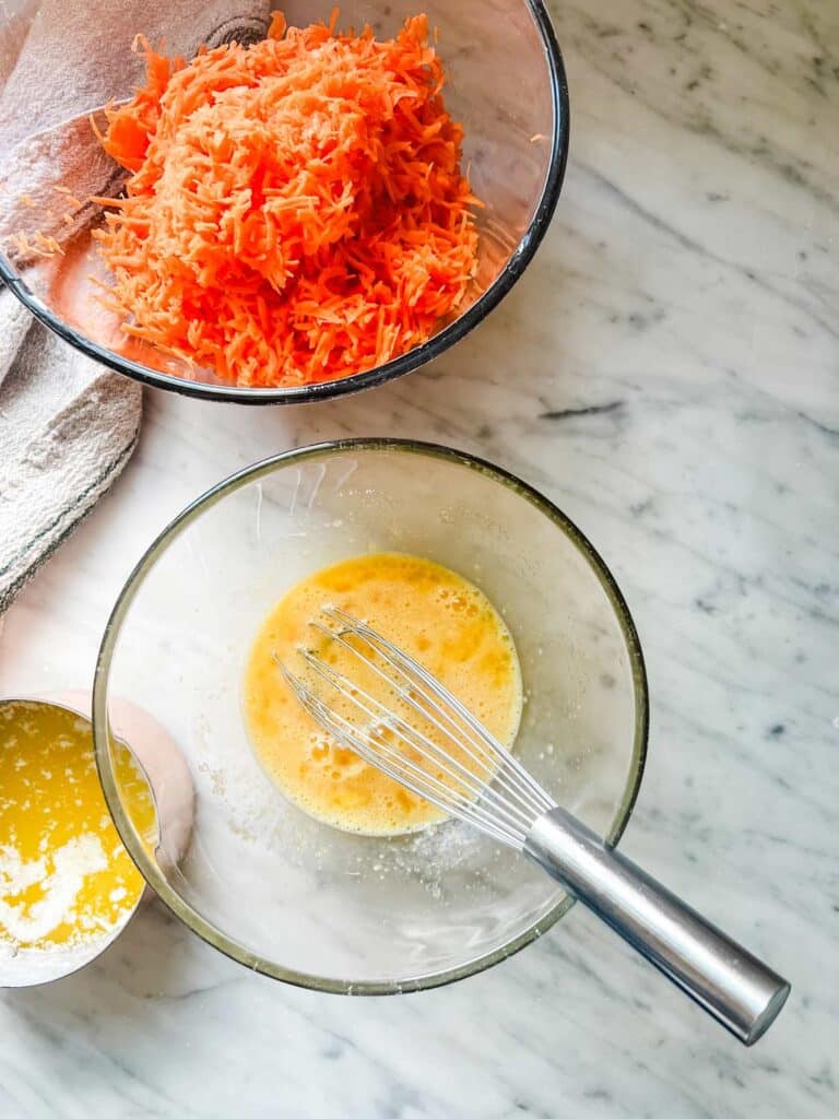 Shredded carrots in one bowl, beaten eggs in another, and melted butter in a ramekin are ready for mixing.