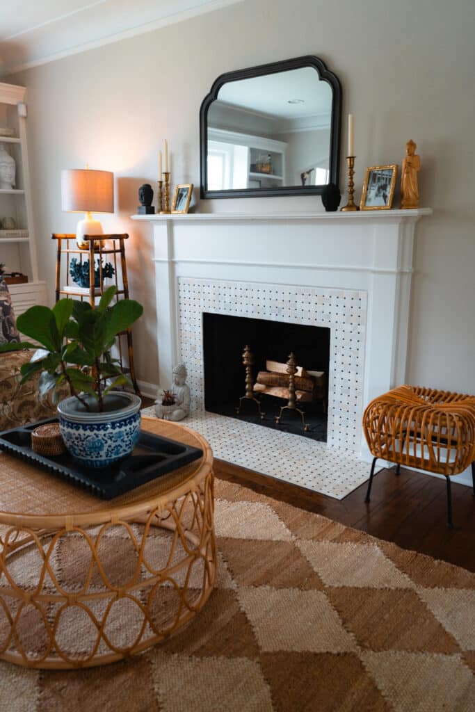 A vintage rattan coffee table, and a rattan stool are next to a white fireplace. On the fireplace mantel rests a vintage mirror.