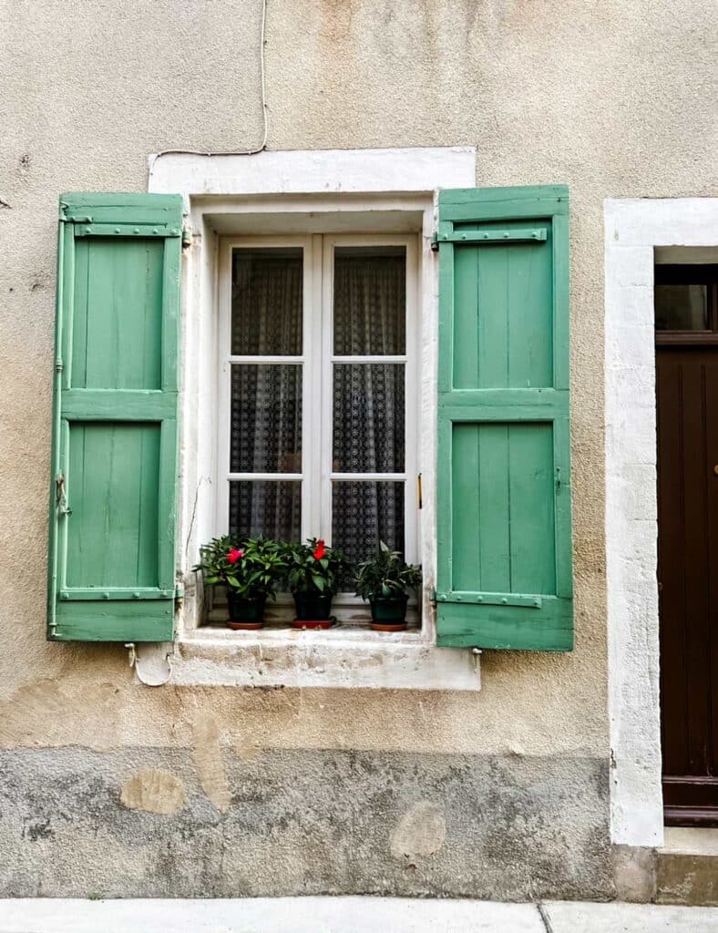Green shutters adorn the window and fresh, potted flowers on the windowsill of a home in Bonnieux, France.