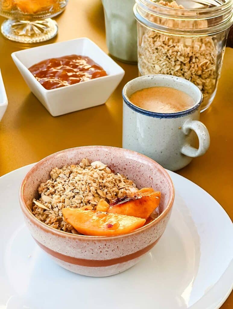 Fresh peaches are served in a bowl of granola. A cup of coffee is also on the table.