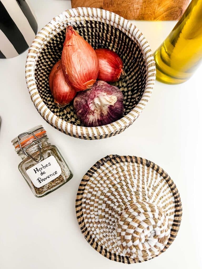 A small basket of shallots and garlic is next to a jar of herbs de Provence.