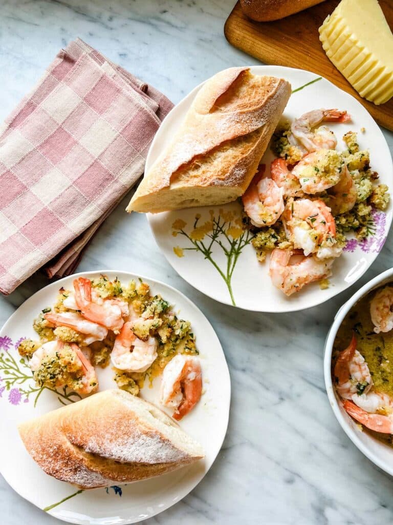 Linen napkins, small plates with flowers, baked shrimp scampi, and a baguette ready to be served.