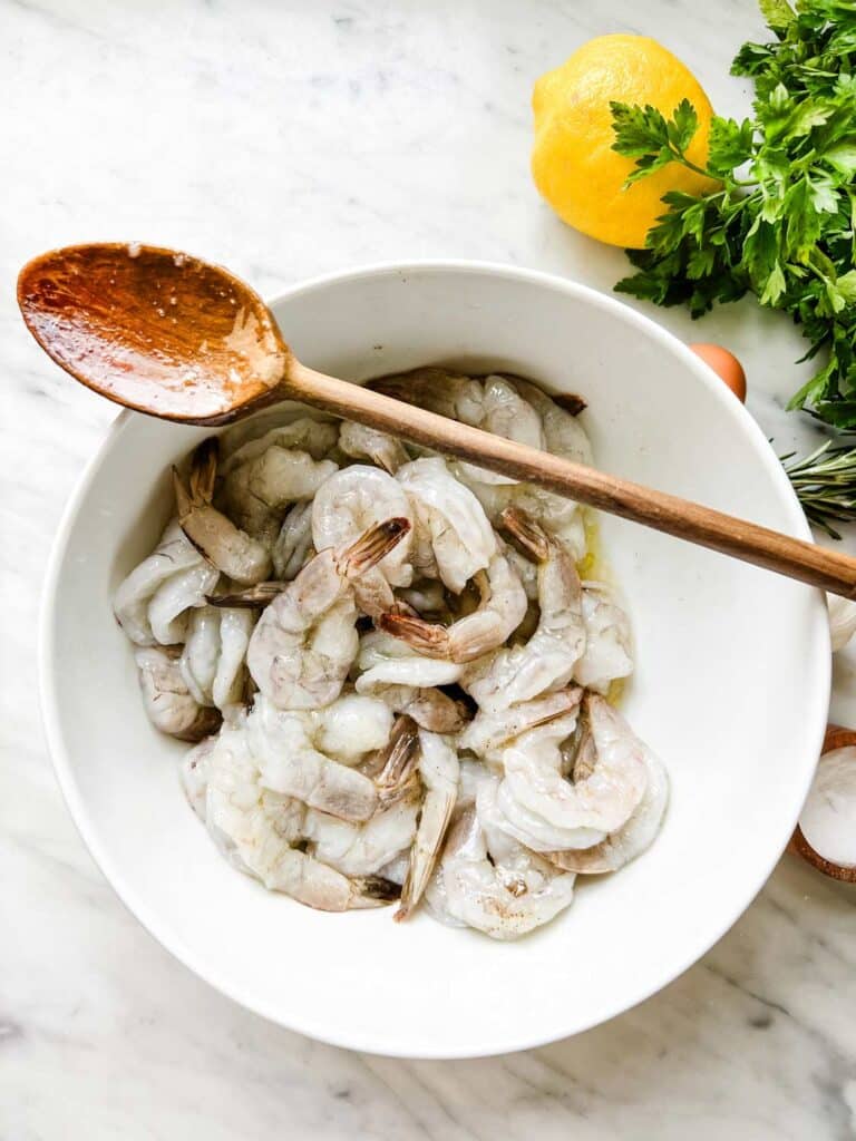 Shrimp is marinating in a white bowl with olive oil, wine, salt and pepper.