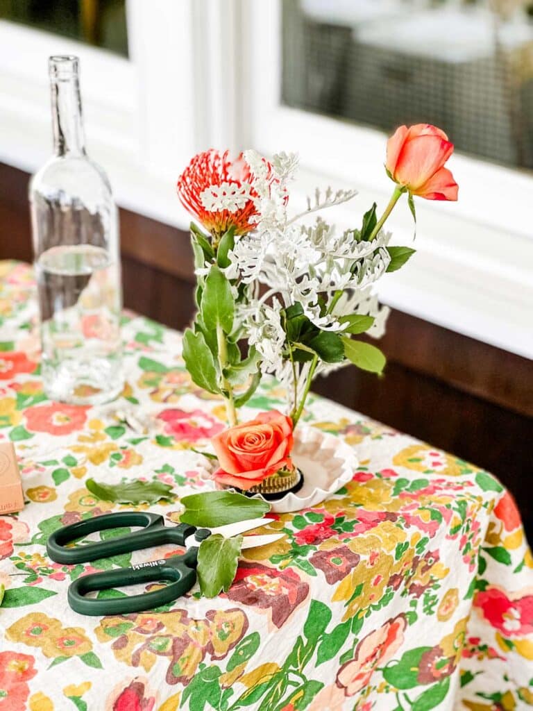 A jar of flowers on a beautiful tablecloth next to a pair of garden clippers.