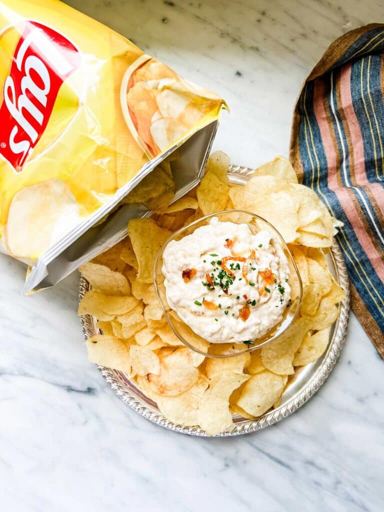 Old fashioned onion dip is served in a small glass bowl on a platter surrounded by classic Lay's potato chips.