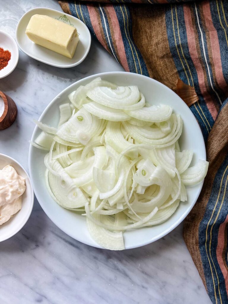Thinly sliced onions are in a white bowl surrounded by ingredients for making old fashioned caramelized onion dip.