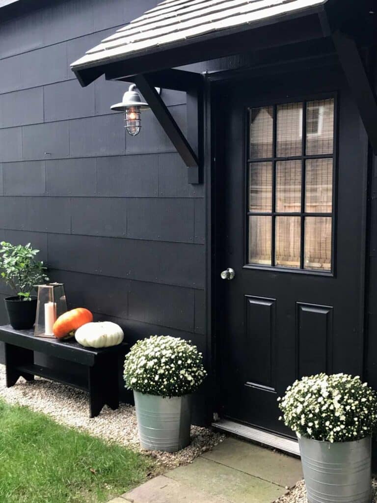 A black garage with a portico over the side door is decorated with plants in galvanized steel containers and a bench with a potted plant, pumpkins, and a candle lantern.