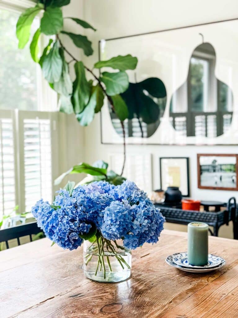 Blue hydrangeas are freshly cut and placed in a glass jar on a long farm table. A candle sits next to them.
