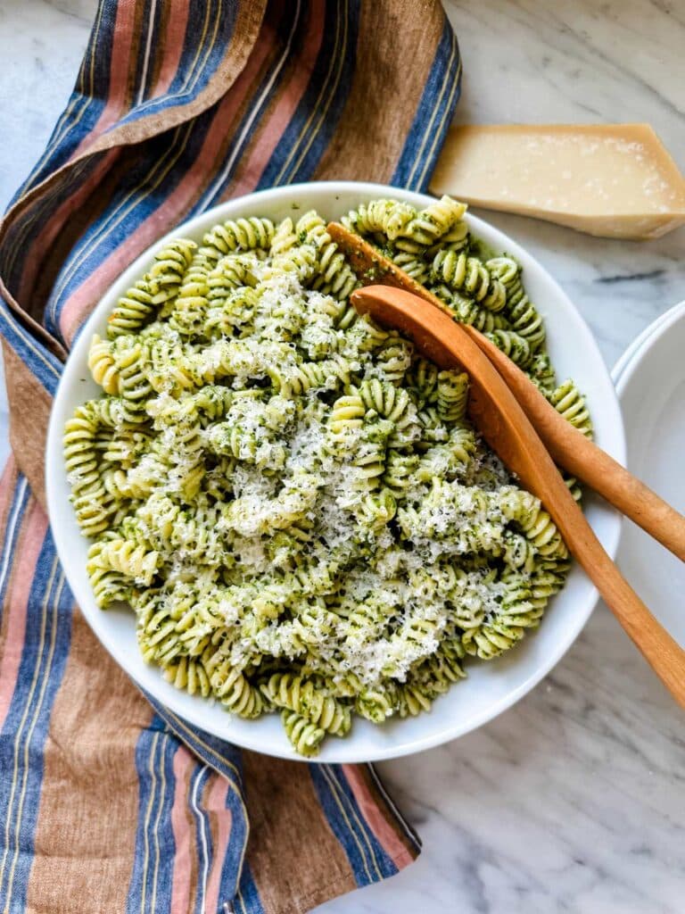 A large white serving bowl with wooden serving spoons is filled with fresh basil pesto pasta.