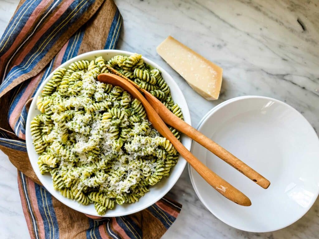 A bowl of freshly made basil pesto pasta in a large white serving bowl with wooden spoons. A block of Parmesan cheese is next to it.