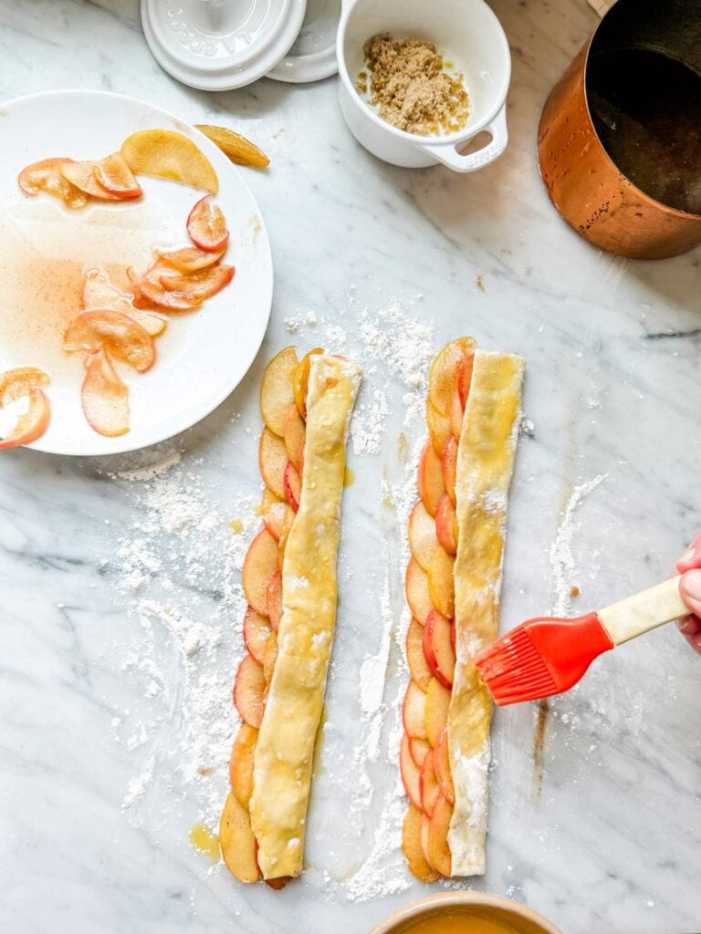 A strip of puff pastry, filled with cooked apples, is being coated with an egg wash.