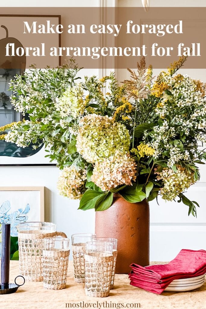 A foraged floral arrangement for fall is in a large crock that is sitting on a vintage farm table. Red napkins are stacked on plates and next to drinking glasses.