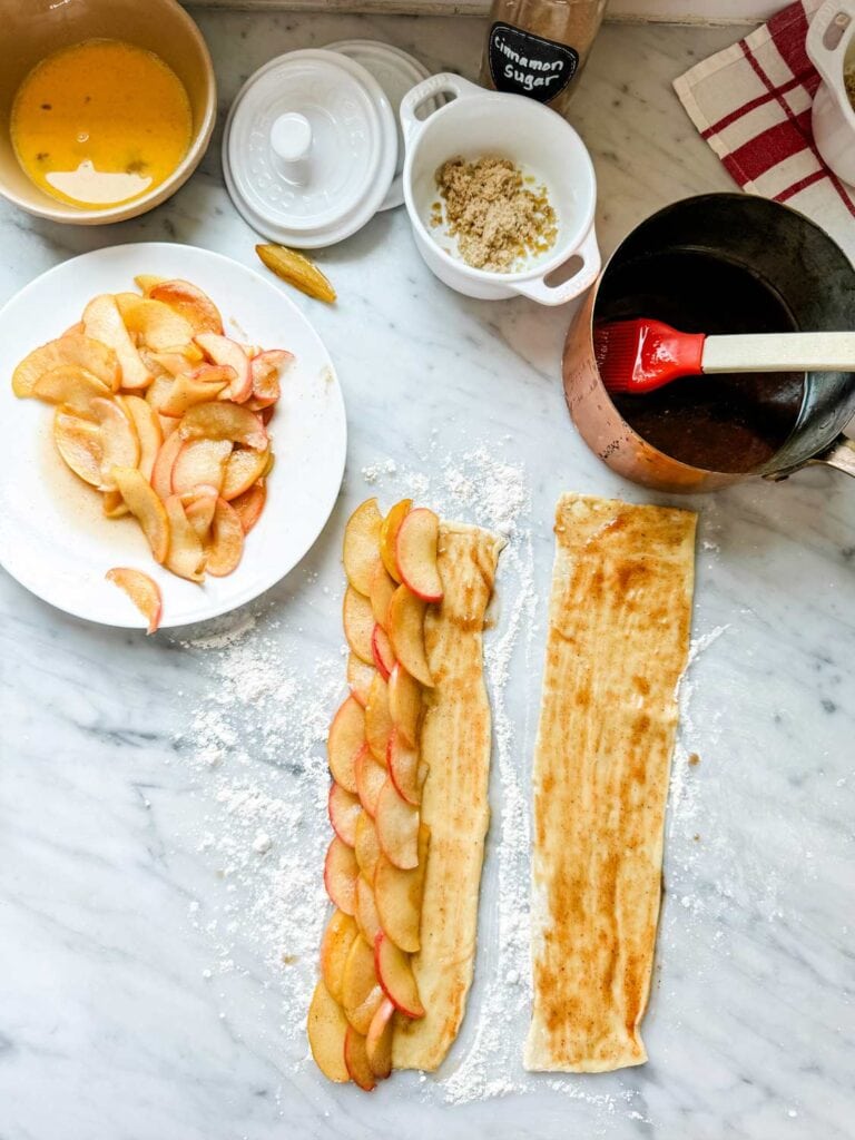 Cooked apple slices have been layered on a strip of puff pastry dough to make apple roses.