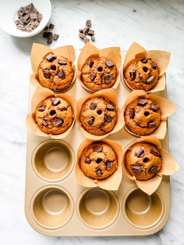 A tray of pumpkin spice muffin with dark chocolate chunks on top and holes in the center of each muffin filled with molten chocolate.