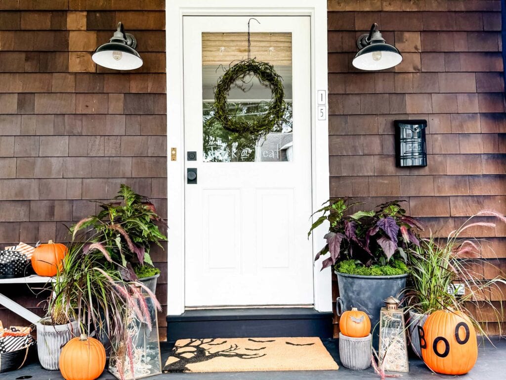 Shingled house white door, wreath, pots woth dark leafy plants, pumpkins. lights on each side of door, lantersn with twinkle lights inside, mailbox on the right of door. 