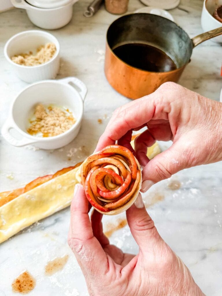 A strip of puff pastry dough is filled with cooked apples and rolled up to form a rose.